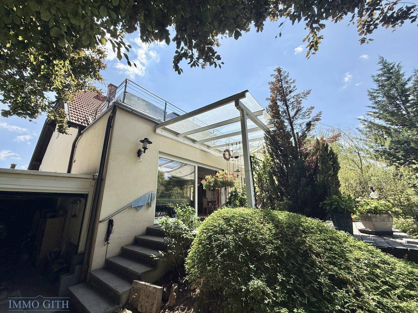 Rückseite des Hauses mit überdachter Terrasse und einer Dachterrasse darüber, umgeben von Grün.