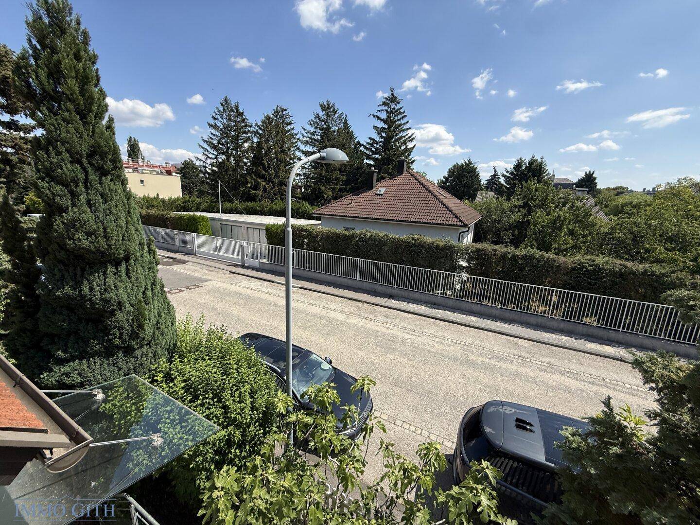 Blick von oben auf eine ruhige Wohnstraße mit gepflegten Hecken und Bäumen unter blauem Himmel.