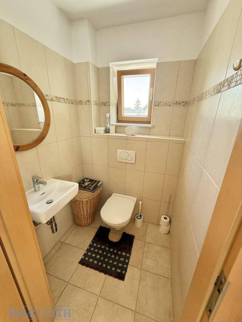 Kleines Gäste-WC mit Fenster, Fliesen an Wänden und Boden sowie einem runden Holzspiegel.