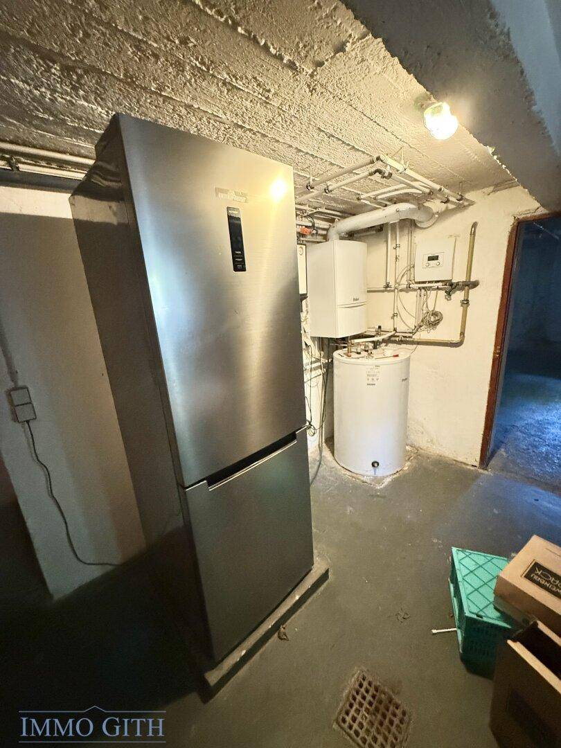 Hauswirtschaftsraum im Keller mit großem Kühlschrank, Warmwasserbereiter und sichtbaren Leitungen.