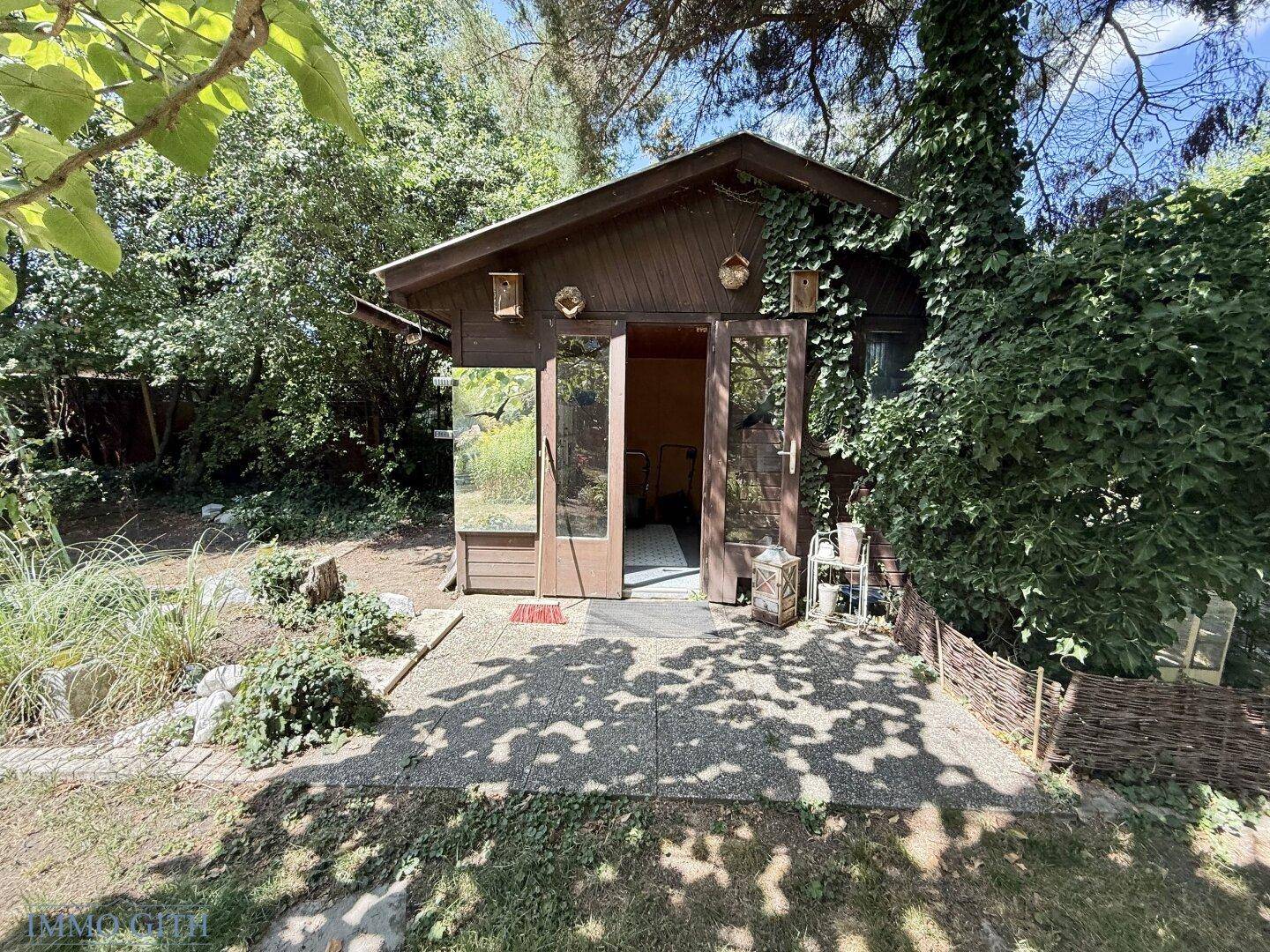 Charmantes Gartenhaus aus Holz mit Glastüren, umgeben von üppigem Grün im Garten.