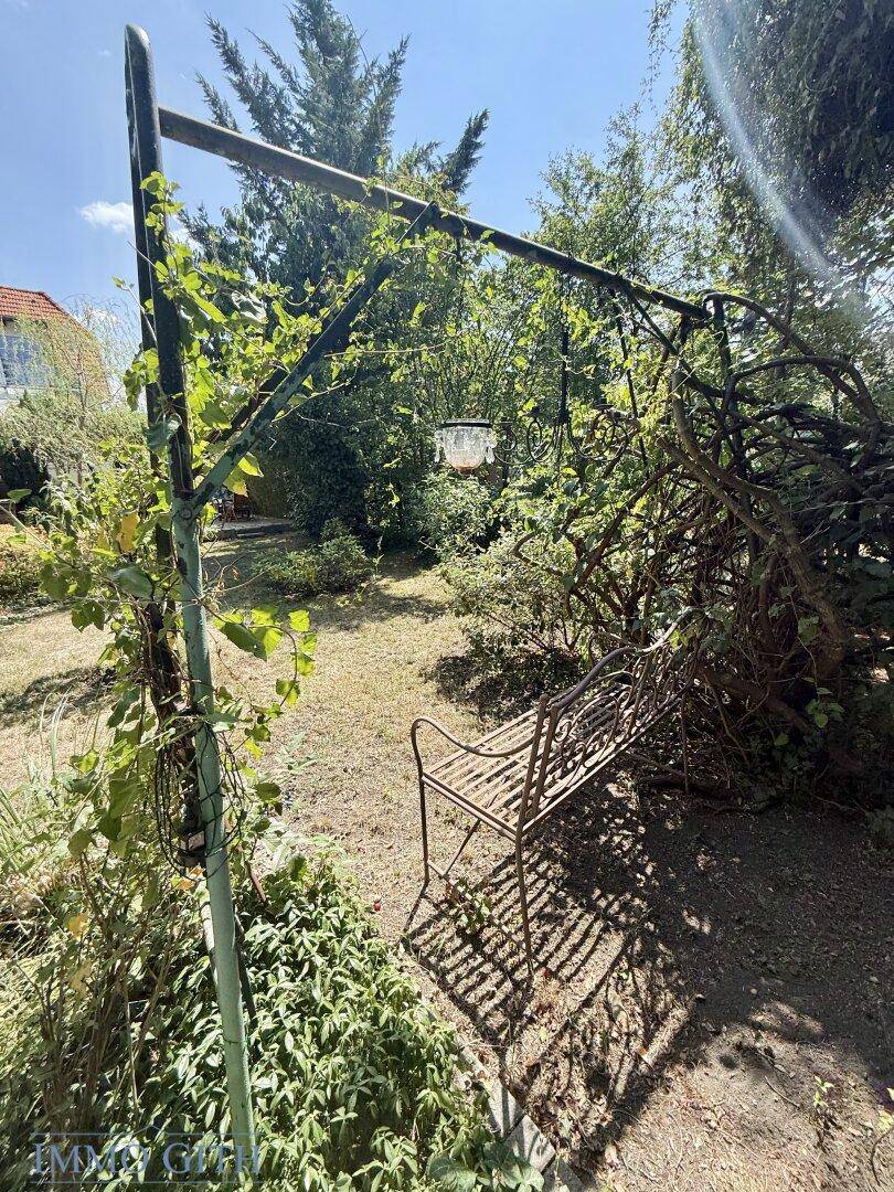 Romantische Gartenbank unter einem bewachsenen Metallbogen, umgeben von üppigem Grün und Bäumen.