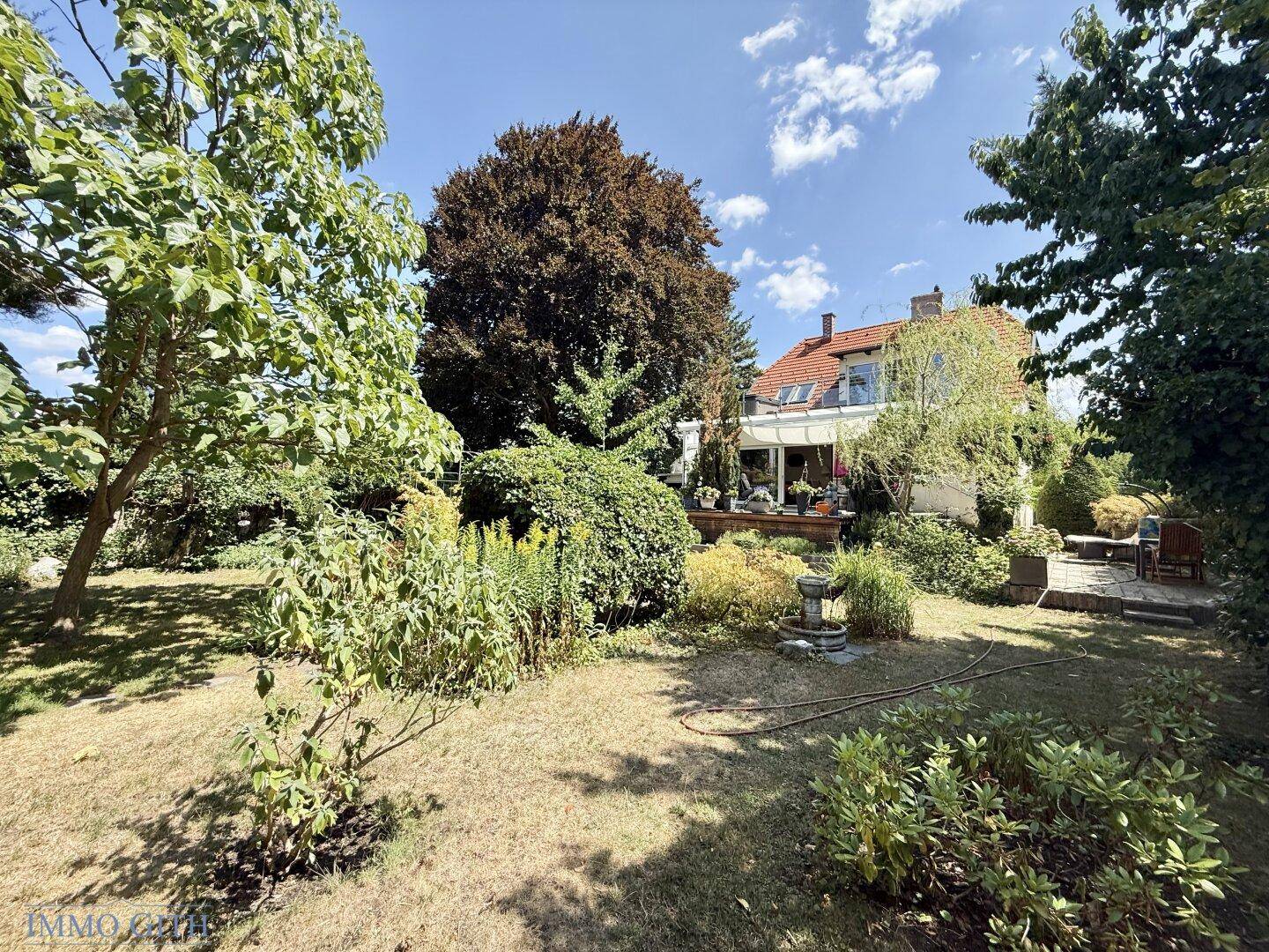 Weitläufiger Garten mit großer Rasenfläche und altem Baumbestand, im Hintergrund das Haus mit Terrasse.