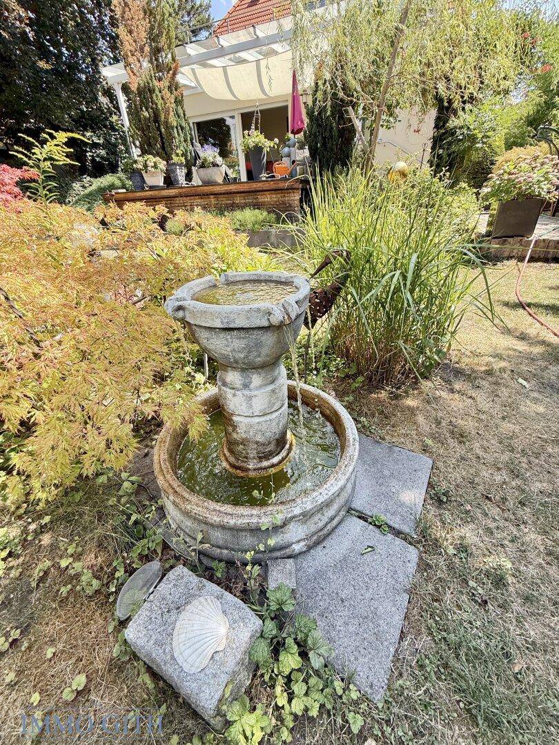 Dekorativer Steingartenbrunnen im grünen Gartenbereich, mit Blick auf die Terrasse des Hauses.