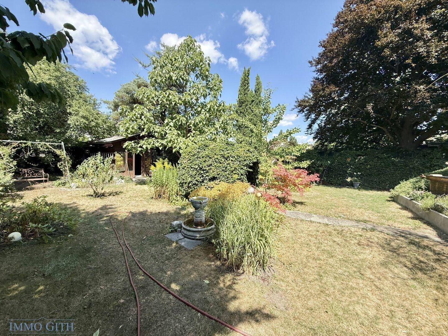 Weitläufiger Garten mit Rasenfläche, dekorativem Brunnen und vielfältigem Baumbestand unter blauem Himmel.