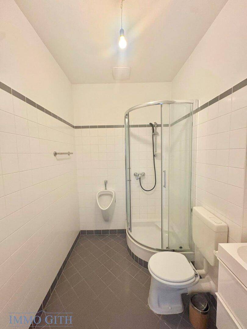 Funktionales Badezimmer mit Dusche, WC und Urinal, gefliest in Weiß und Grau.