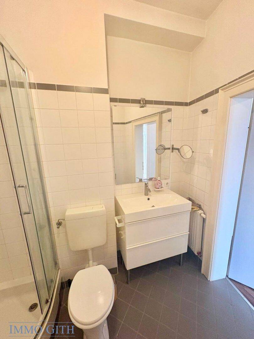 Badezimmer mit Dusche, WC, Waschbecken und Spiegel, gefliest in Weiß und Grau.