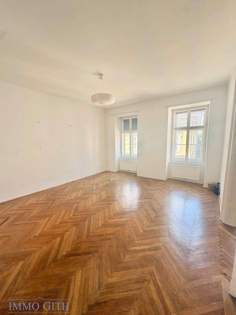 Helles Zimmer mit Fischgrätparkett, hohen Decken und zwei Fenstern.