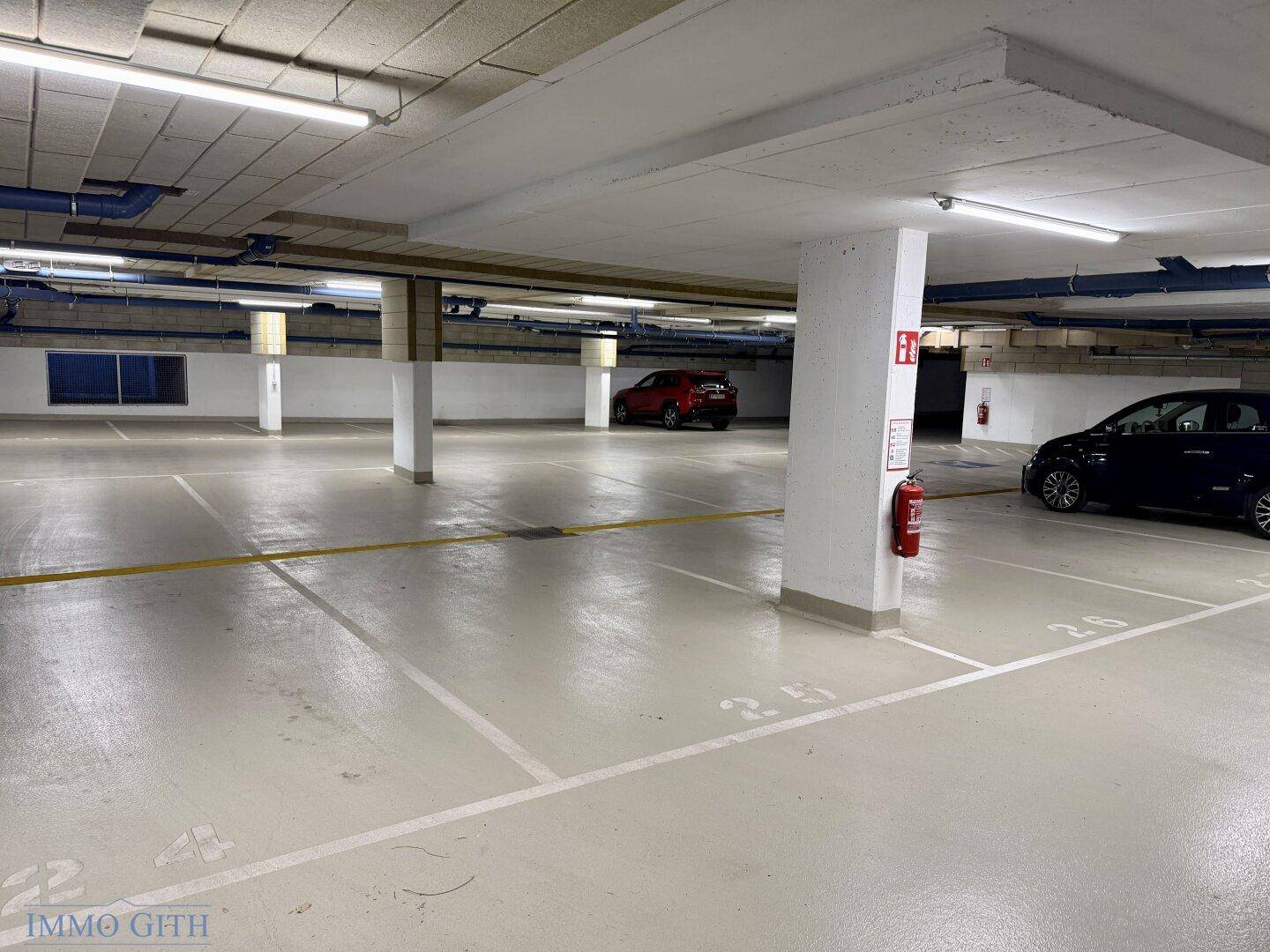 Helle Tiefgarage mit nummerierten Parkplätzen, Säulen und einem roten Feuerlöscher.