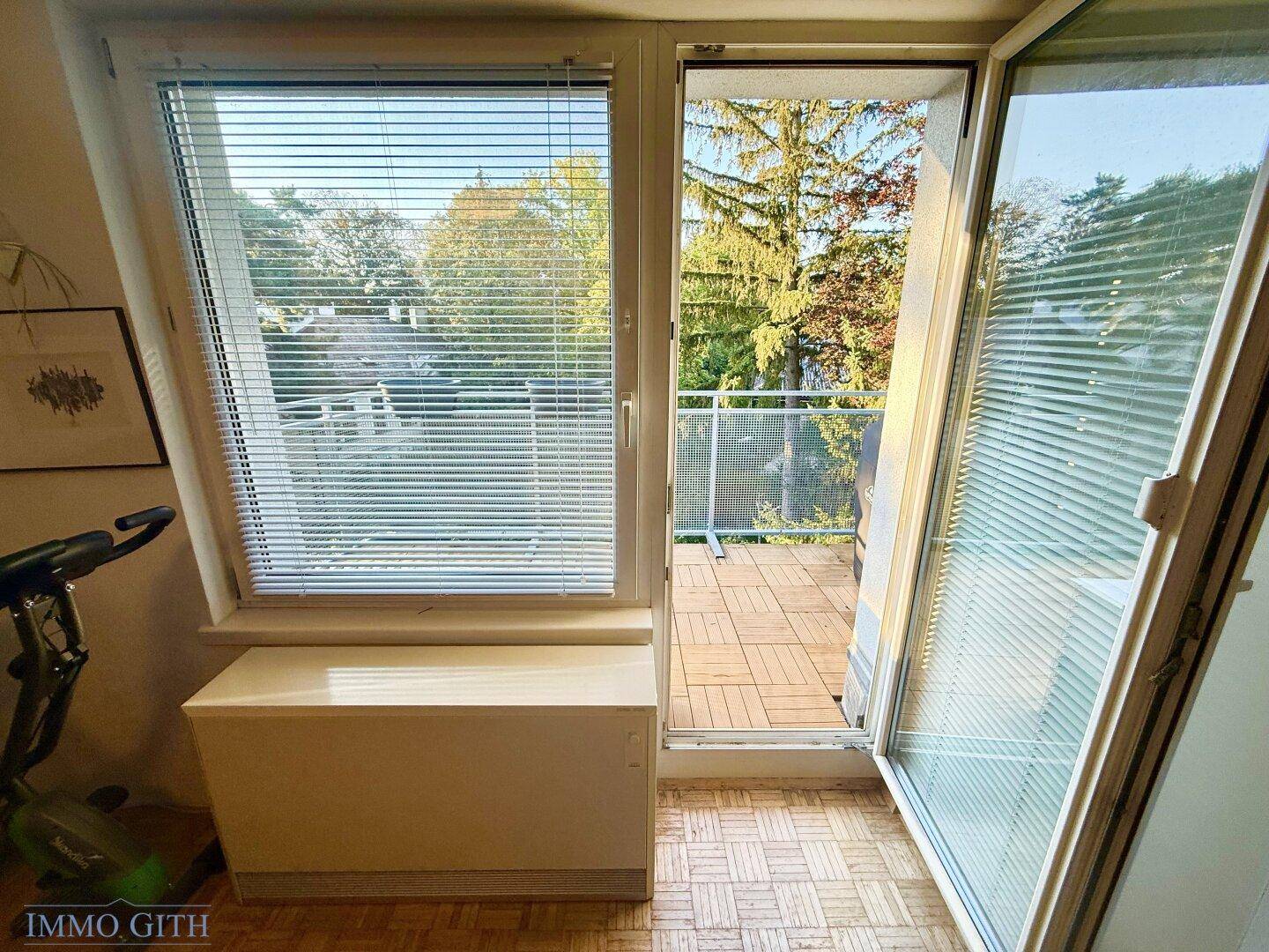 Blick vom Schlafzimmer auf den Balkon mit Holzboden und grüner Umgebung.