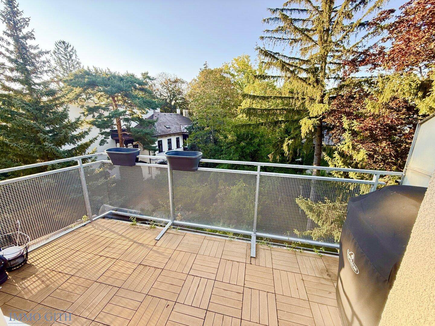 Sonniger Balkon mit Holzdielenboden, Metallgeländer und Blick ins Grüne.