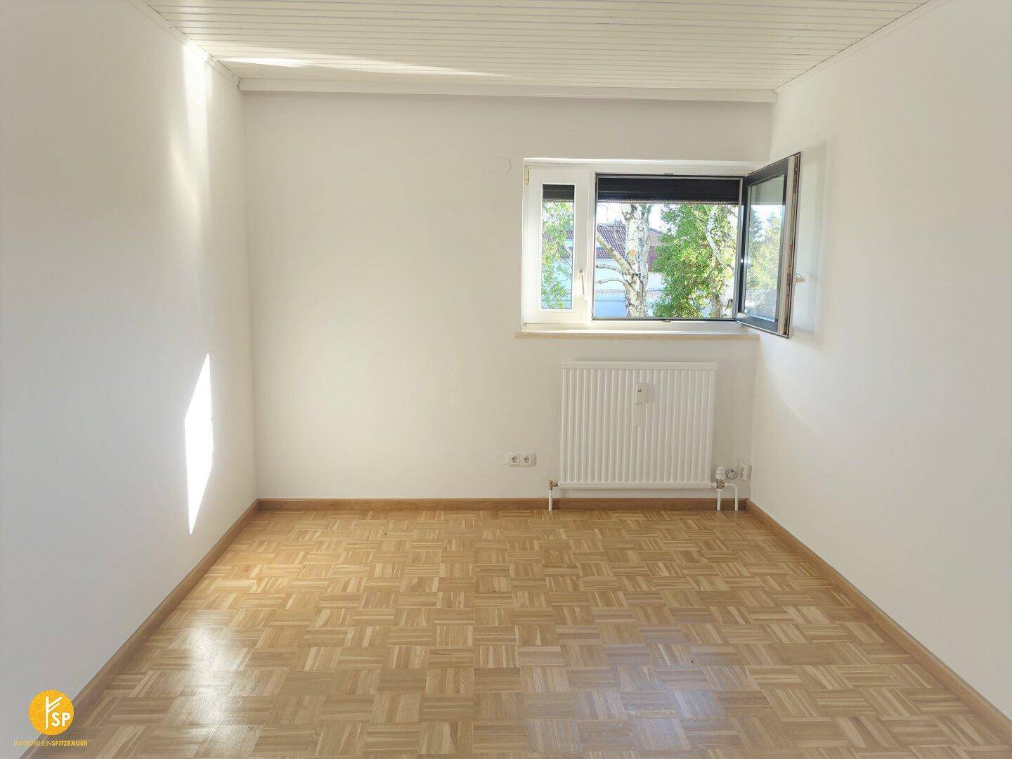 Helles Zimmer mit Parkettboden, weißen Wänden und Fenster mit Blick ins Grüne.