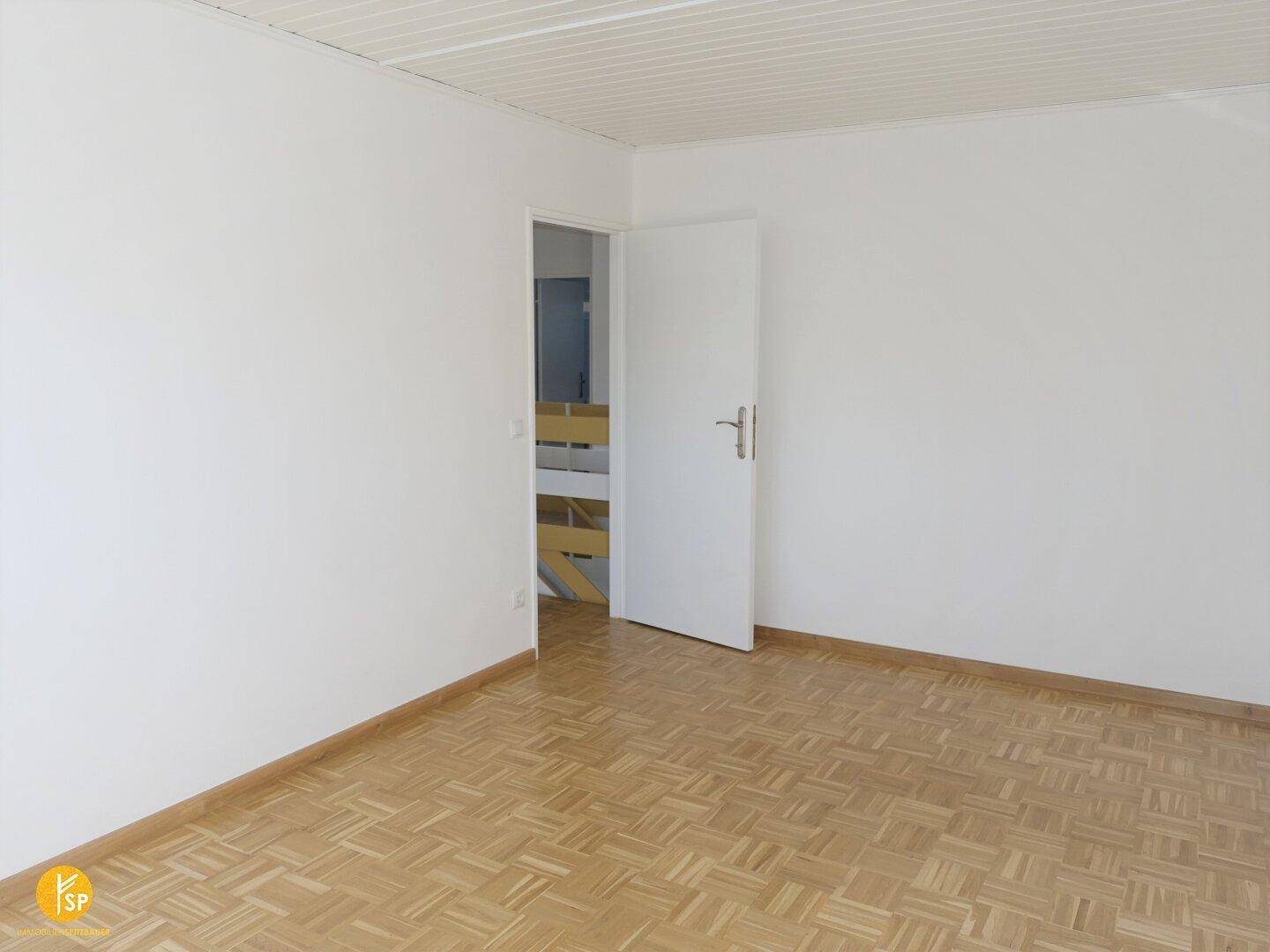 Geräumiges Zimmer mit Parkettboden und weiß gestrichenen Wänden, Tür offen.