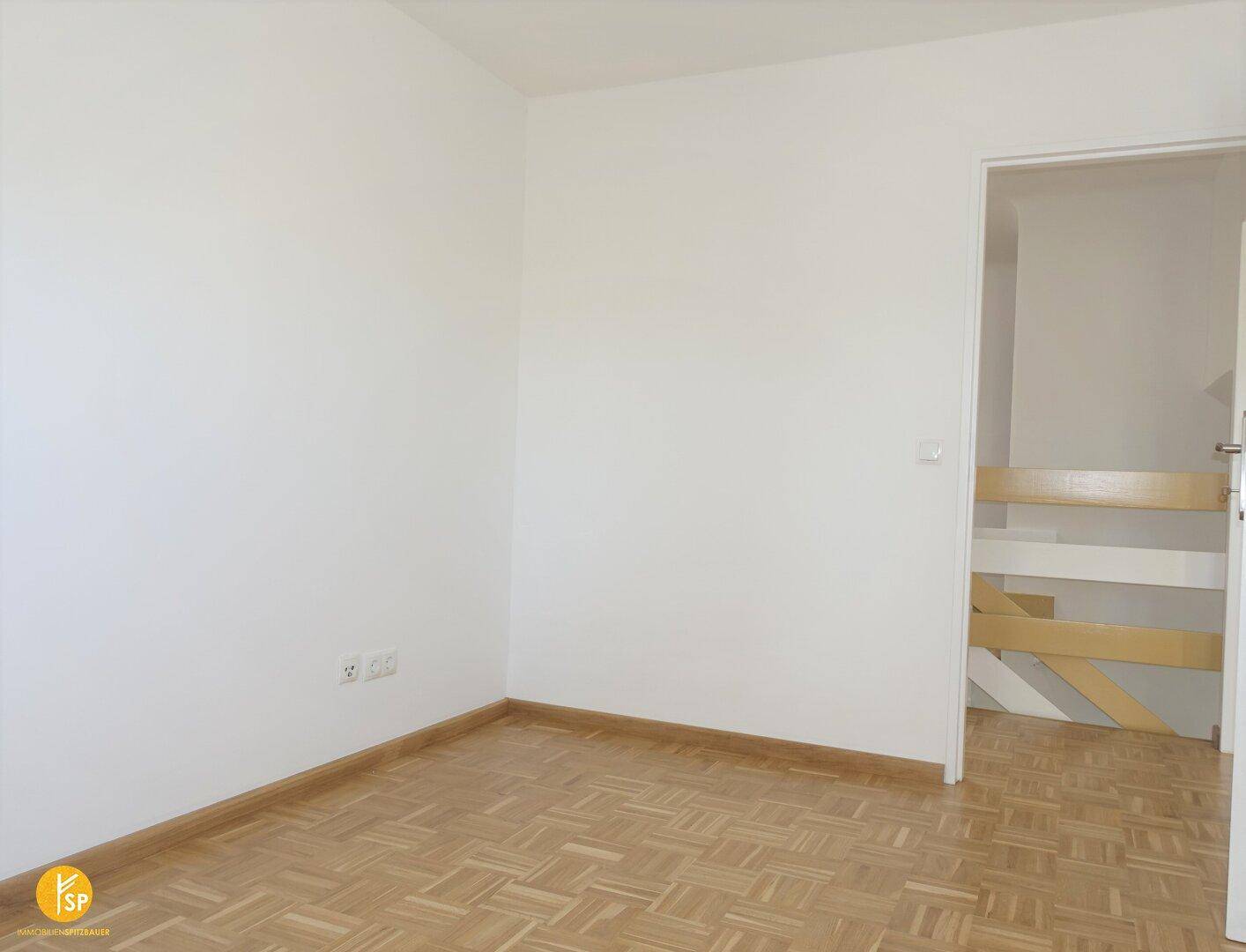Schlichtes Zimmer mit Parkettboden und weiß gestrichenen Wänden, Tür offen.