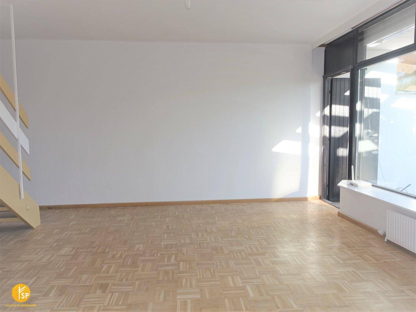 Geräumiges Wohnzimmer mit Parkettboden, großen Fenstern und Zugang zu einem Außenbereich.