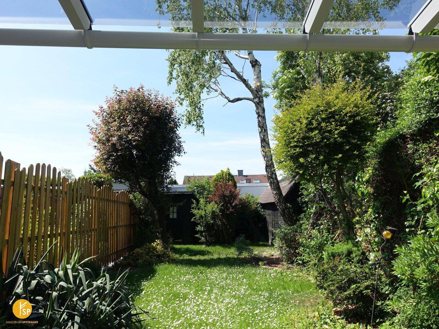 Sonniger Garten mit Rasenfläche und üppiger Bepflanzung unter einer Terrassenüberdachung.