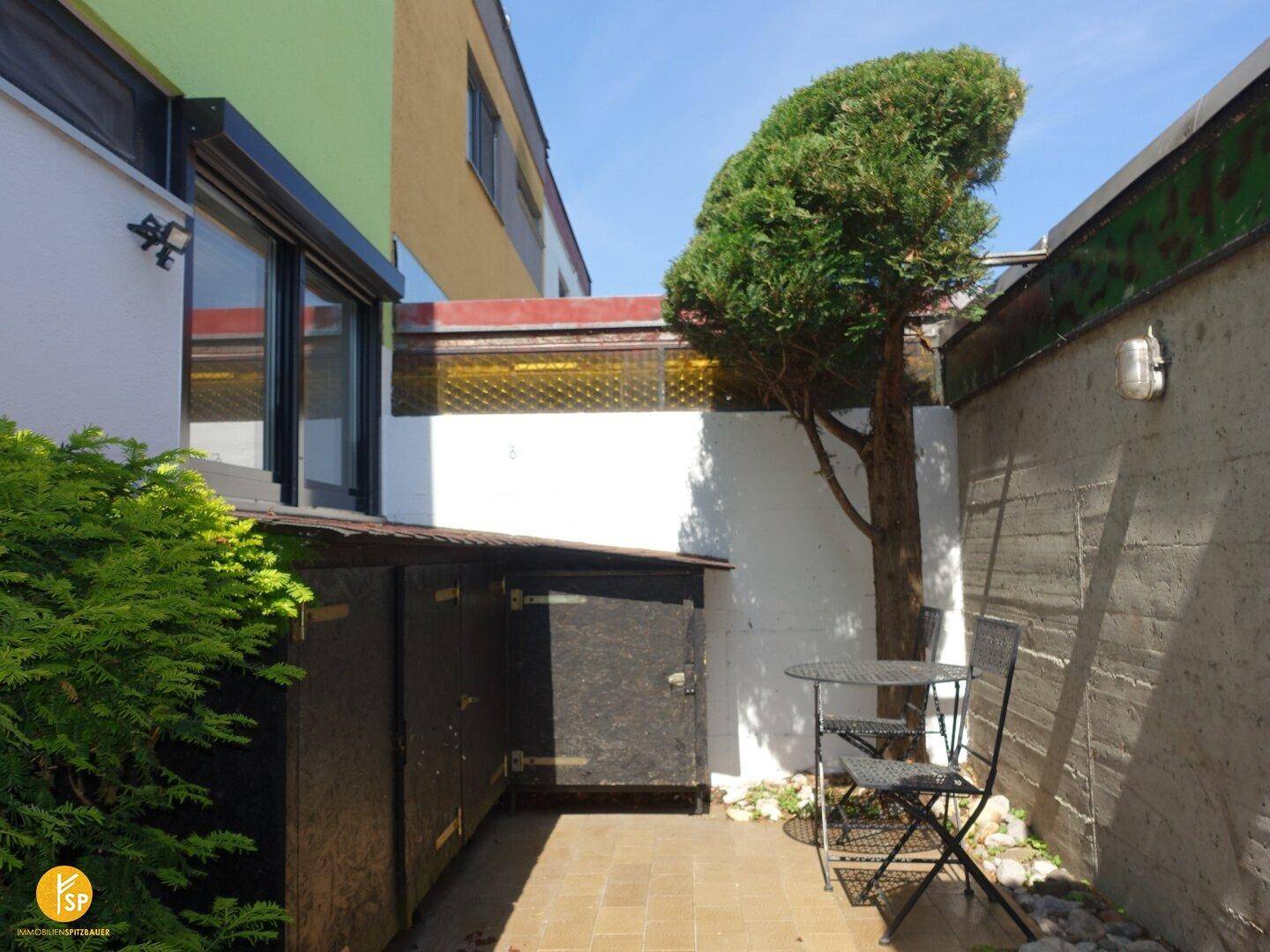 Kleiner, sonniger Patio mit Sitzgelegenheiten, umgeben von Pflanzen und Abstellschuppen.