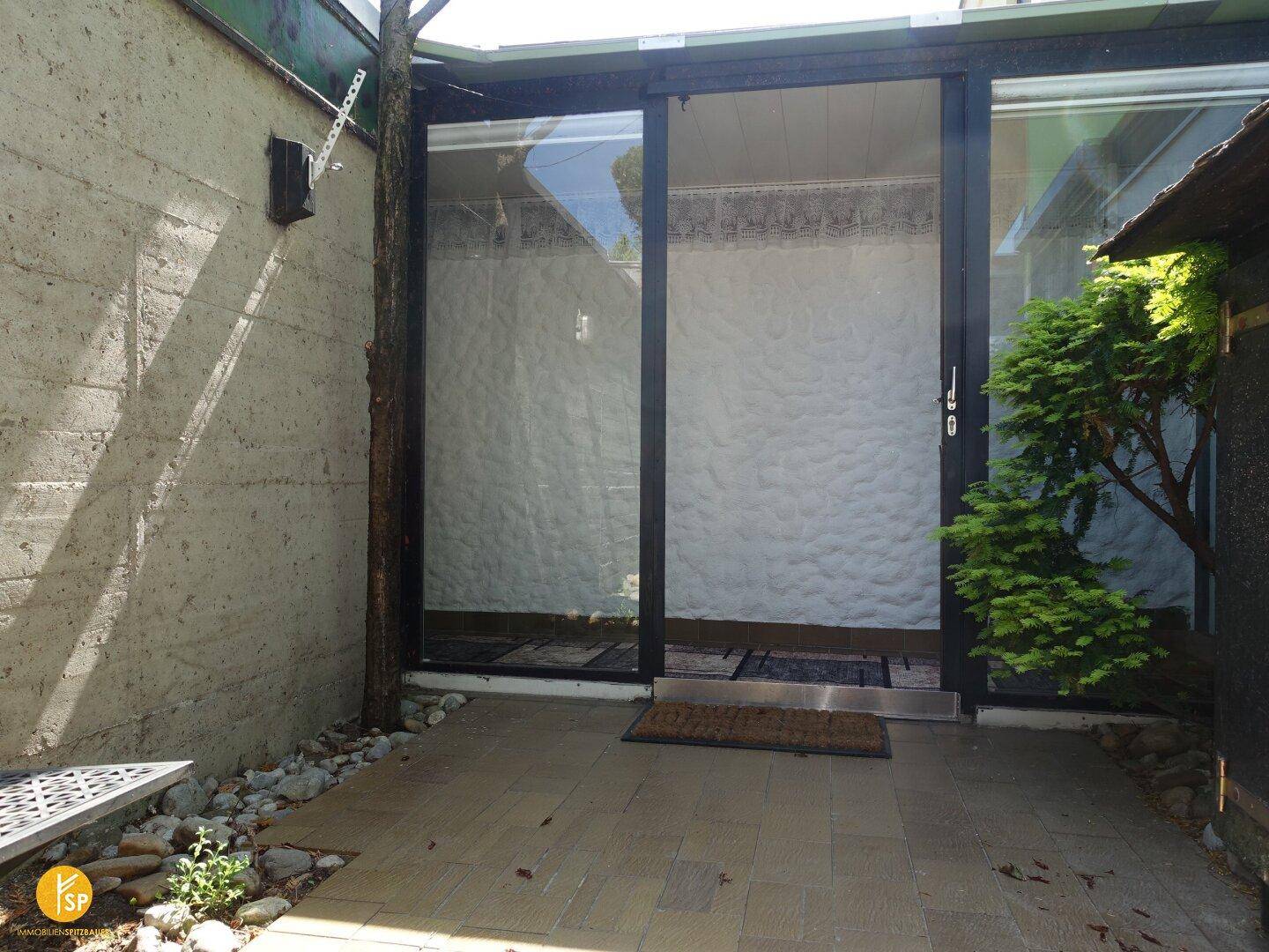 Gepflasterter Außenbereich mit Betonwand und großen Glasfronten zum Gebäude.