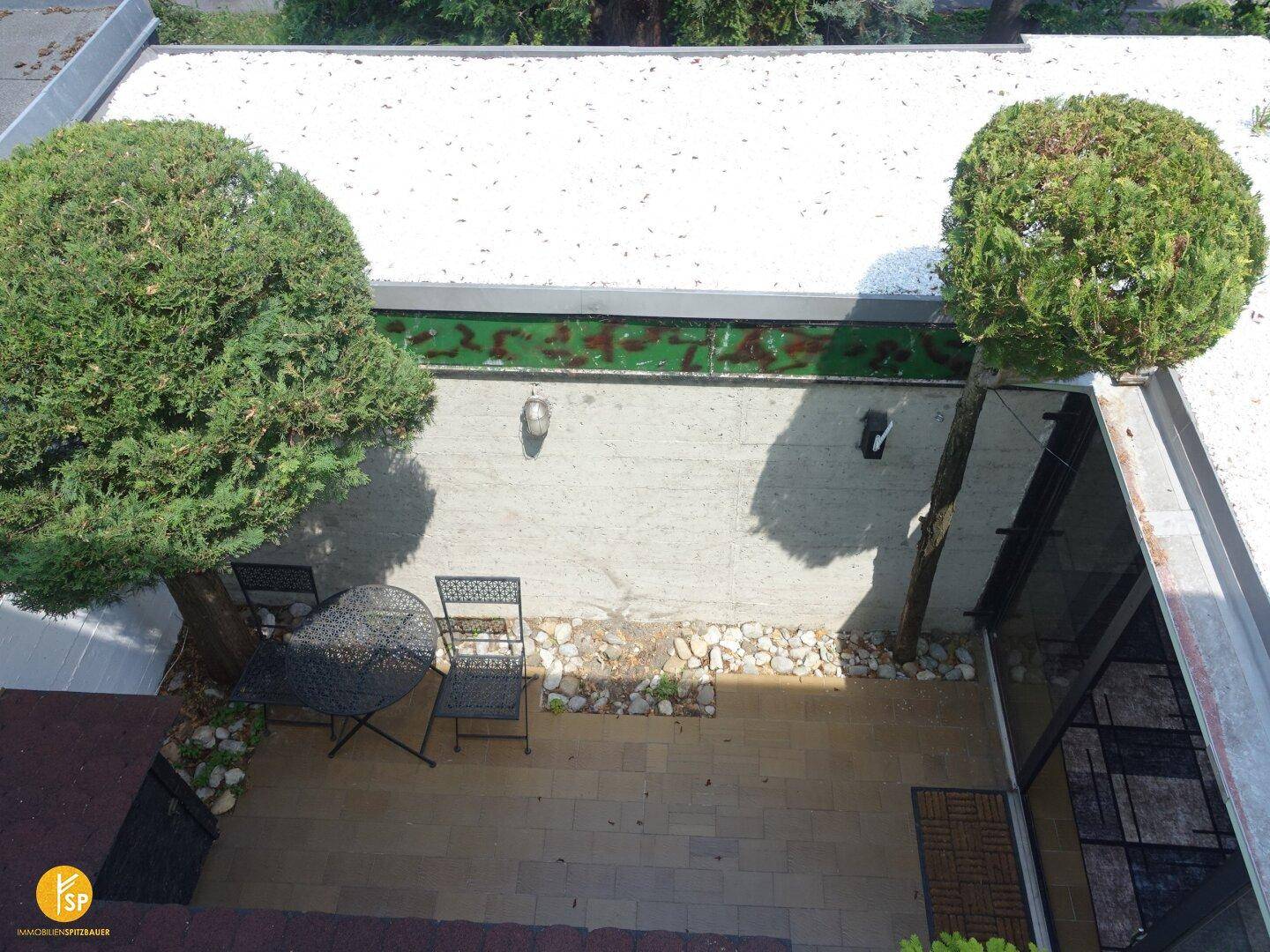 Blick von oben auf eine kleine gepflasterte Terrasse mit Sitzgelegenheiten und Bäumen.