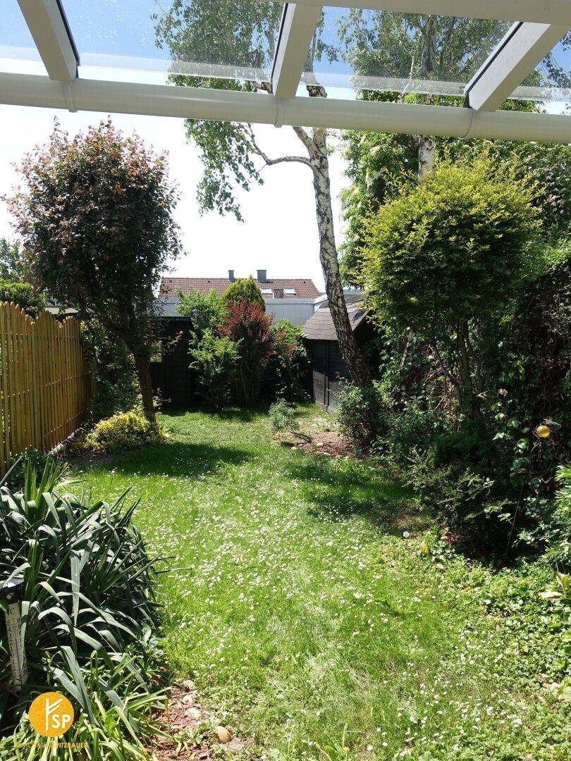 Gepflegter Gartenbereich mit Rasenfläche und verschiedenen Pflanzen, von einer überdachten Terrasse aus gesehen.