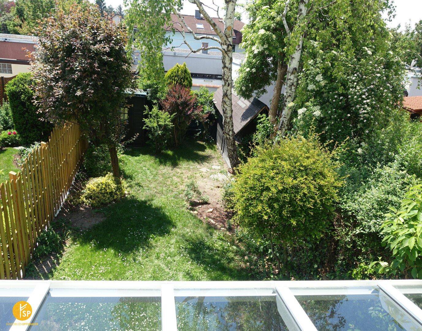 Sonniger Blick von oben auf den Garten mit Rasen, Bäumen und Holzzaun.