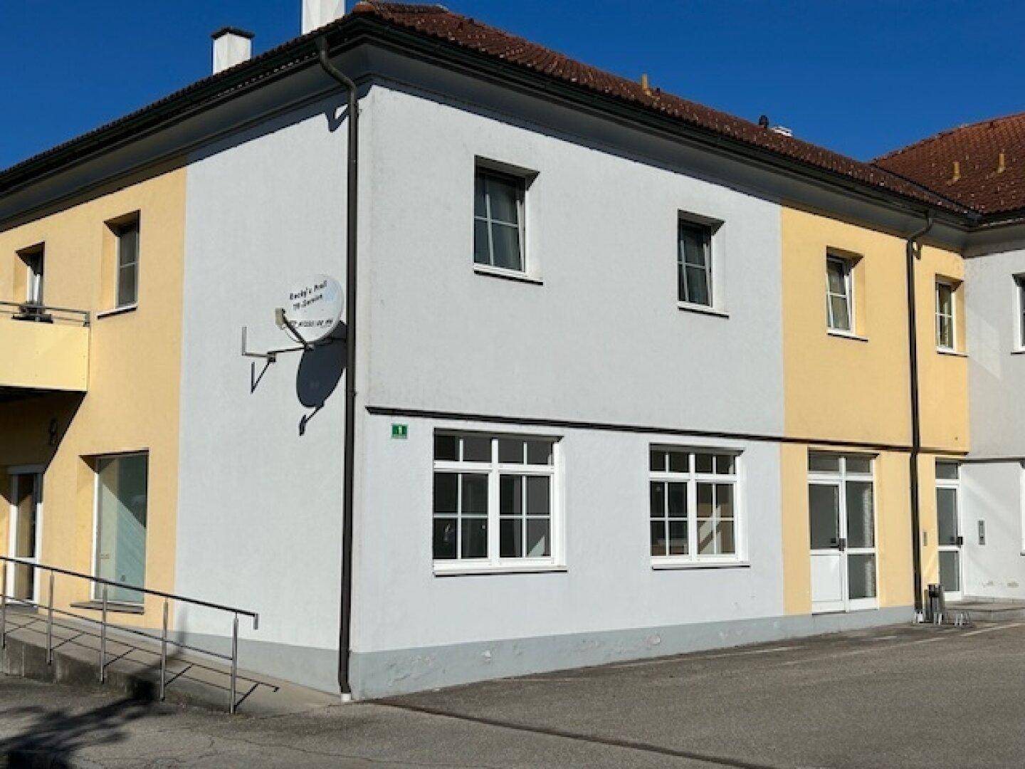 Seitenansicht eines Gebäudes mit hellgrauer und gelber Fassade, einem Ziegeldach und einer Zugangsrampe.