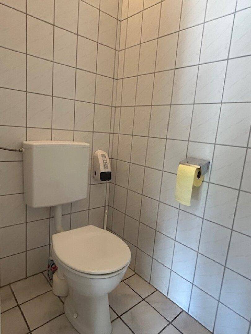 Einfache Toilette mit weißen Wand- und Bodenfliesen, ausgestattet mit einem WC und Toilettenpapierhalter.