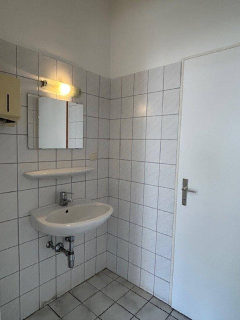 Badezimmer mit weißen Wand- und Bodenfliesen, einem Waschbecken, Spiegel und Beleuchtung über dem Spiegel.