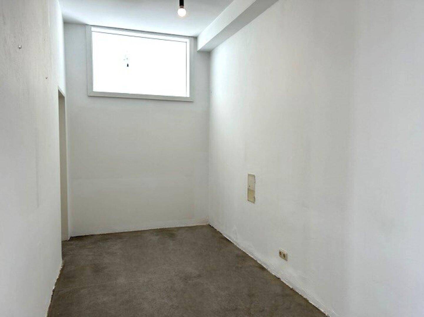 Kompakter, heller Raum mit einem hohen Fenster und Betonboden, geeignet als Abstellraum oder kleines Büro.