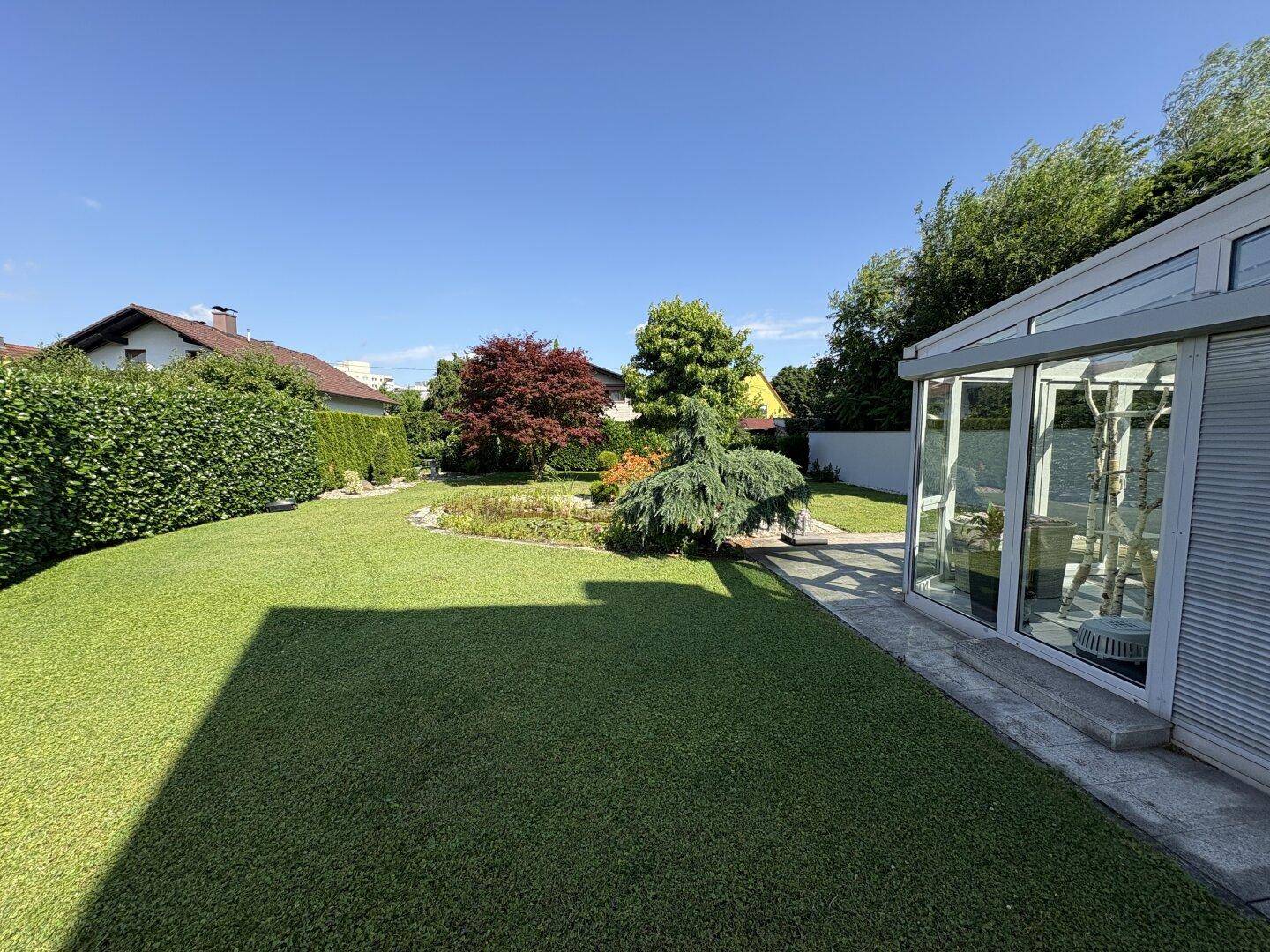 Weitläufiger grüner Rasen mit Bäumen und einem modernen Wintergarten am Haus.
