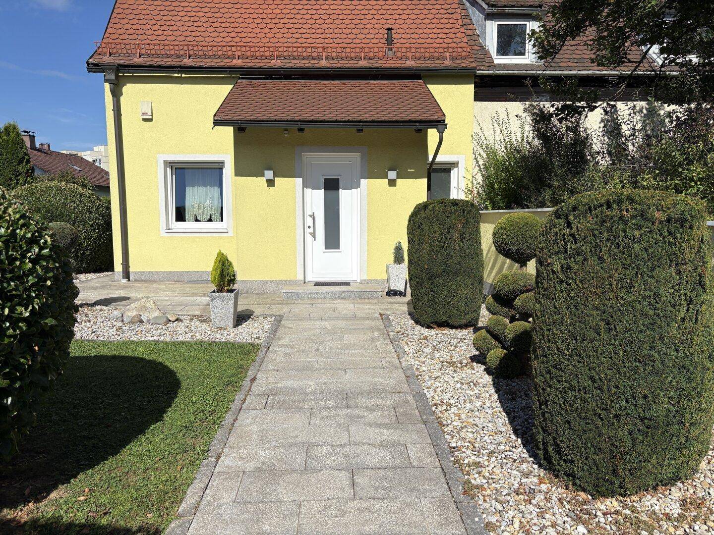 Eingangsbereich eines gelben Hauses mit gepflastertem Weg und grünen Büschen im Vorgarten.