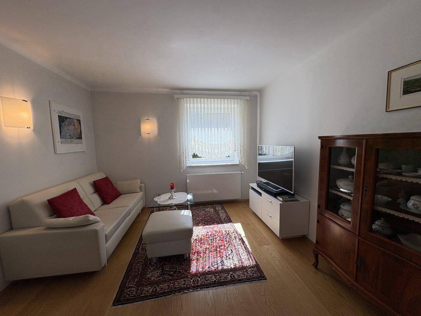 Helles Wohnzimmer mit Sofa, Teppich und Holzschrank, durchflutet von natürlichem Licht.
