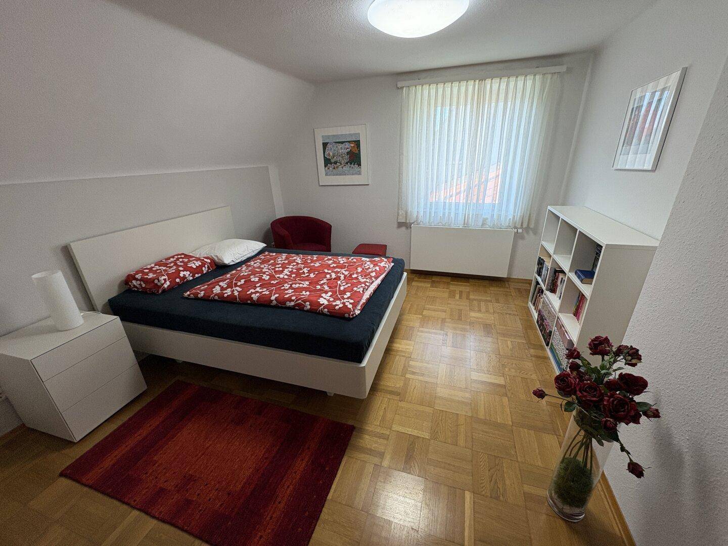 Helles Schlafzimmer mit Parkettboden, großem Bett und Fenster mit Vorhängen.