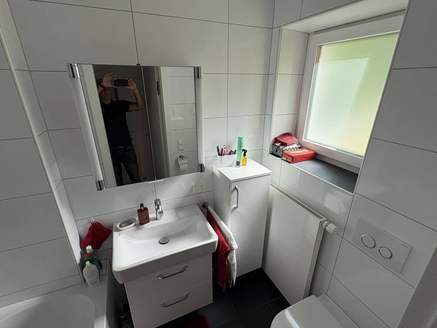 Helles Badezimmer mit weißen Fliesen, Fenster, Waschbecken und Toilette.