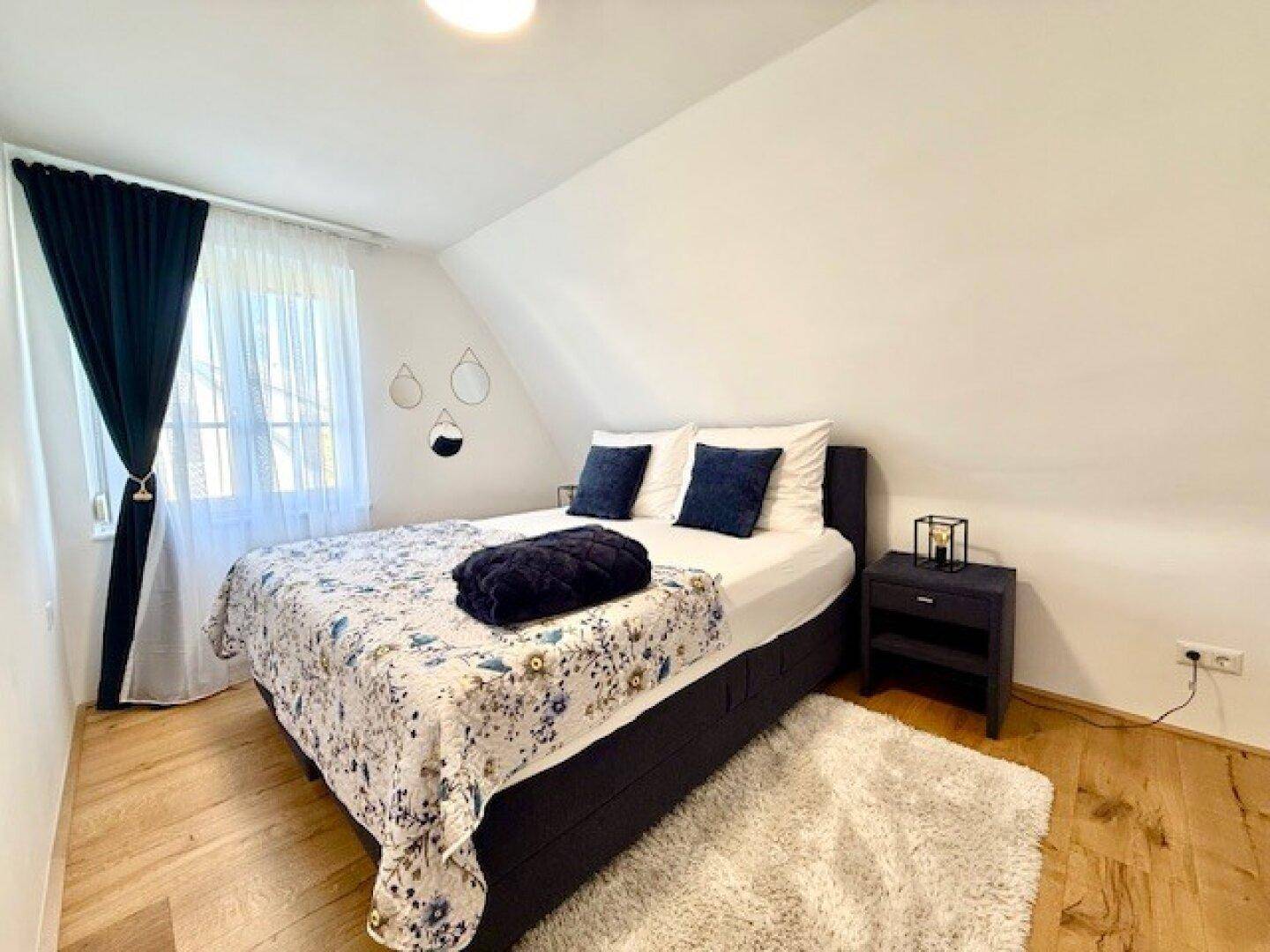 Schlafzimmer mit Holzboden, Dachschräge und gemütlichem Bett mit dunklen Kissen und Decke.
