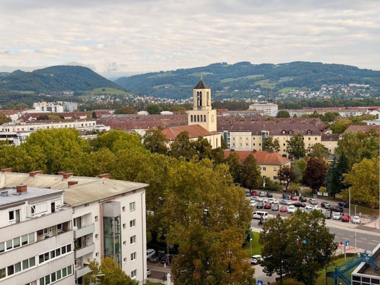 Weitläufiger Blick über die Stadt mit grünen Hügeln und einem Kirchturm im Hintergrund.