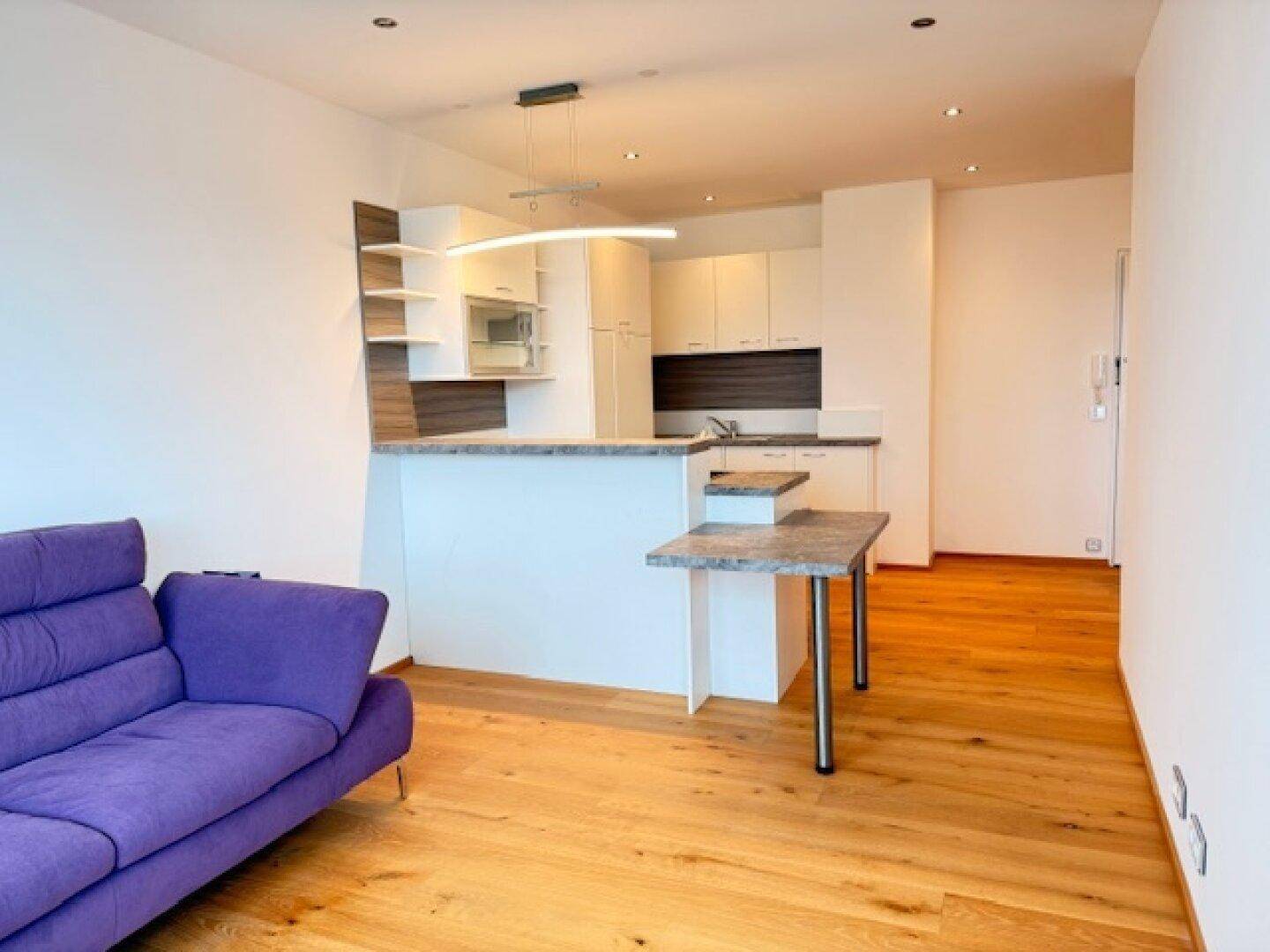 Offener Wohnbereich mit violetter Couch, moderner Küche und hellem Holzparkettboden.