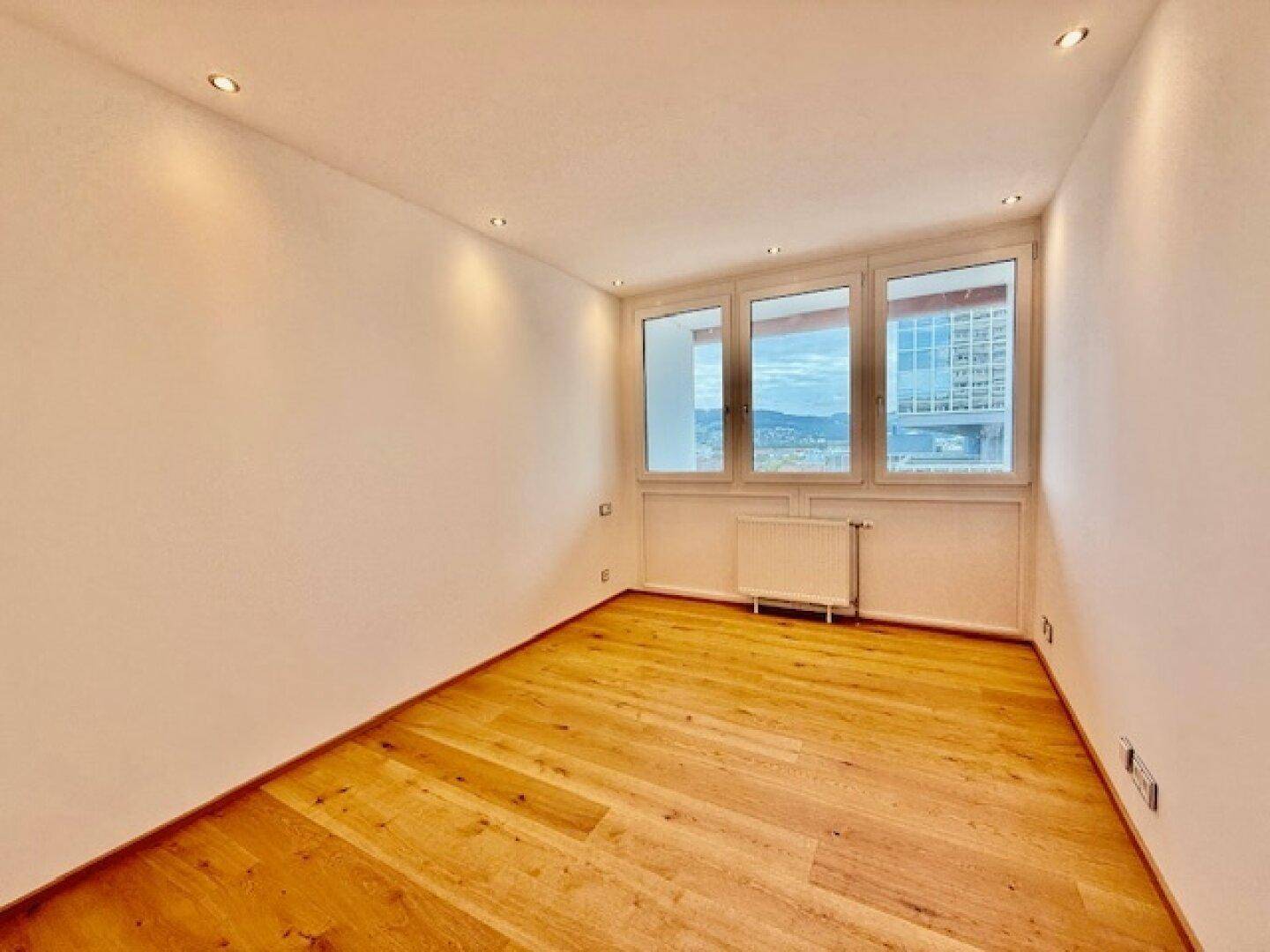 Heller, leerer Raum mit Holzfußboden und großen Fenstern, die einen Stadtblick bieten.