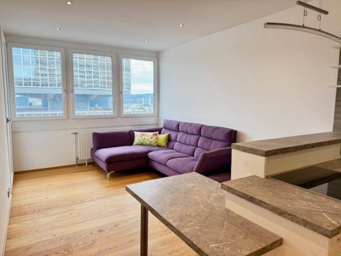 Helles Wohnzimmer mit violetter Eckcouch, großen Fenstern und weitem Stadtblick.