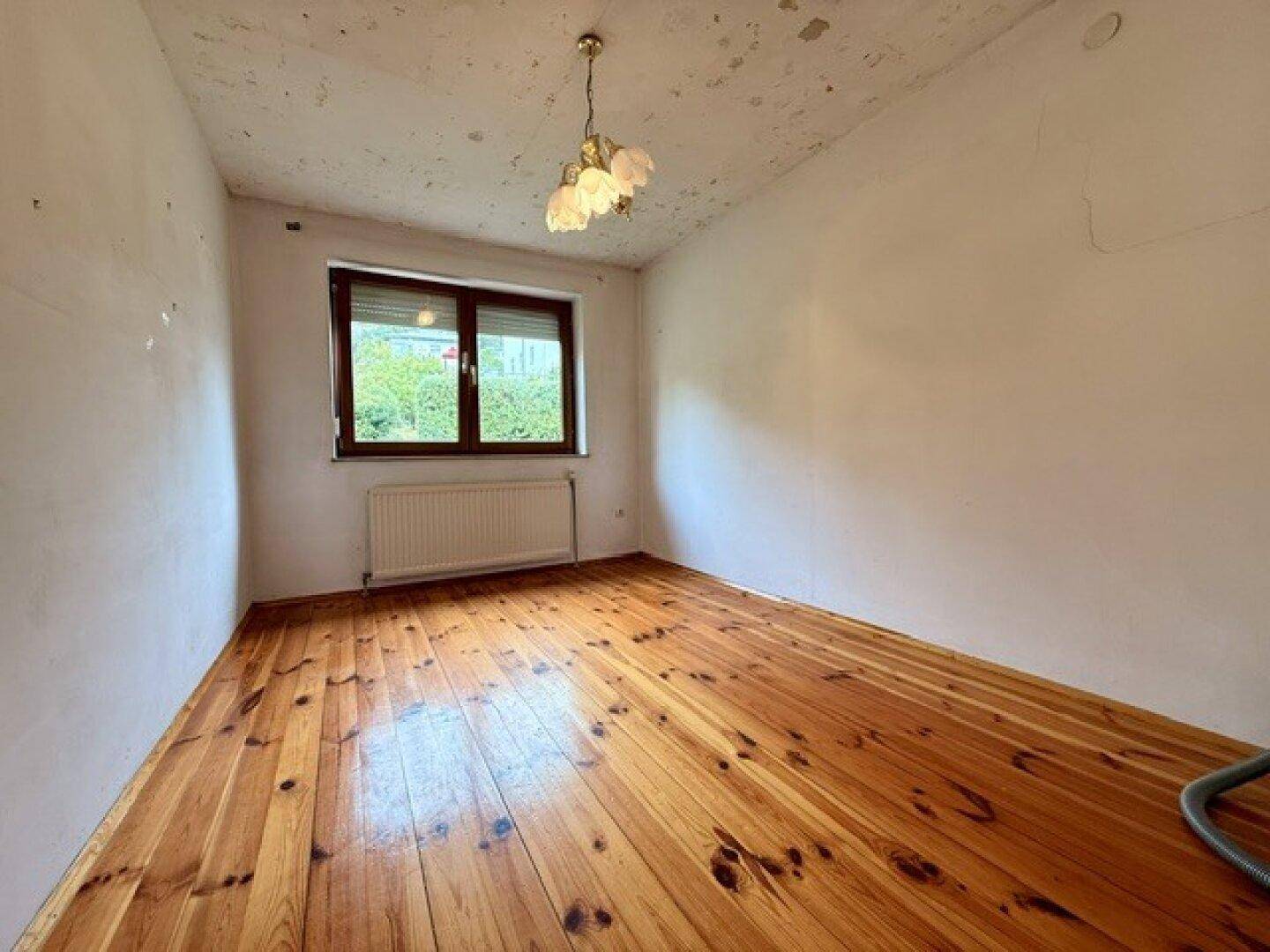 Helles Zimmer mit Holzdielenboden, Fenster und Heizkörper, bereit zur Renovierung.