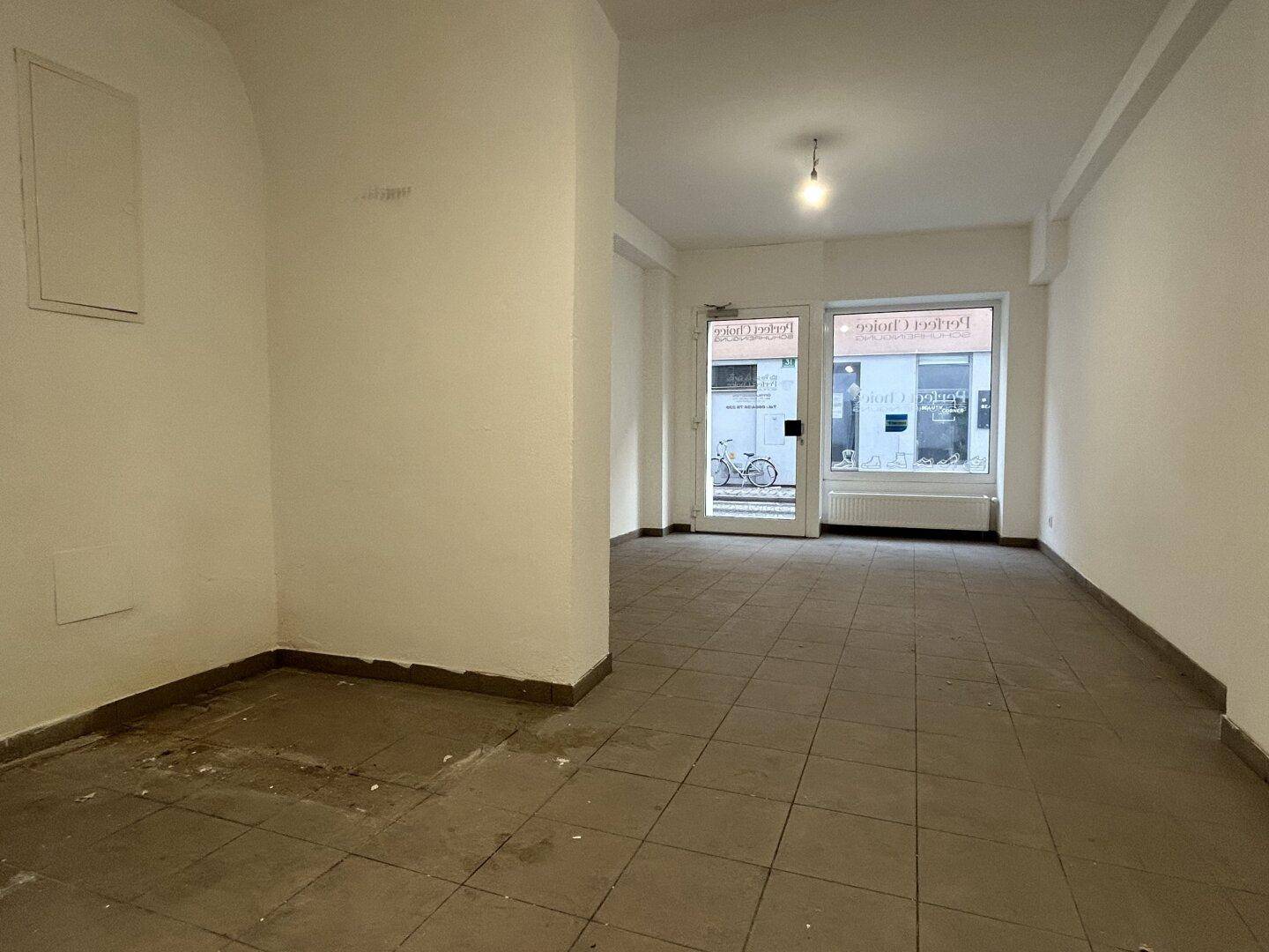 Geräumiger, leerer Gewerberaum mit Fliesenboden und großen Schaufenstern zur Straße hin.