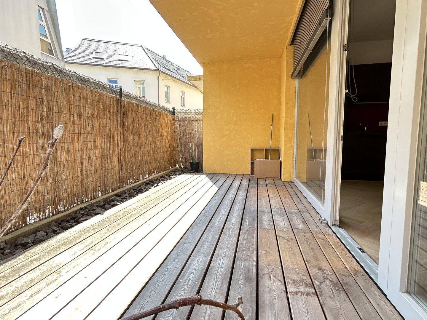 Sonniger Terrassenbereich mit Holzdeck, Bambuszaun und gelber Fassade des Gebäudes.