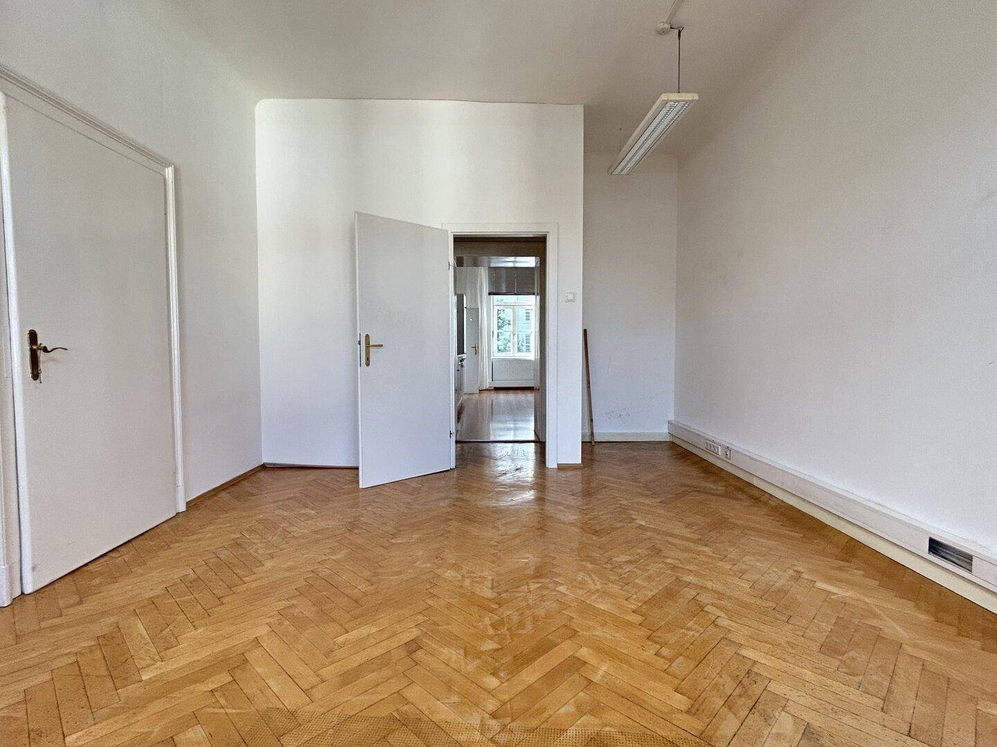 Leerer Raum mit klassischem Fischgrätparkett und einer offenen Tür zu einem angrenzenden Zimmer.