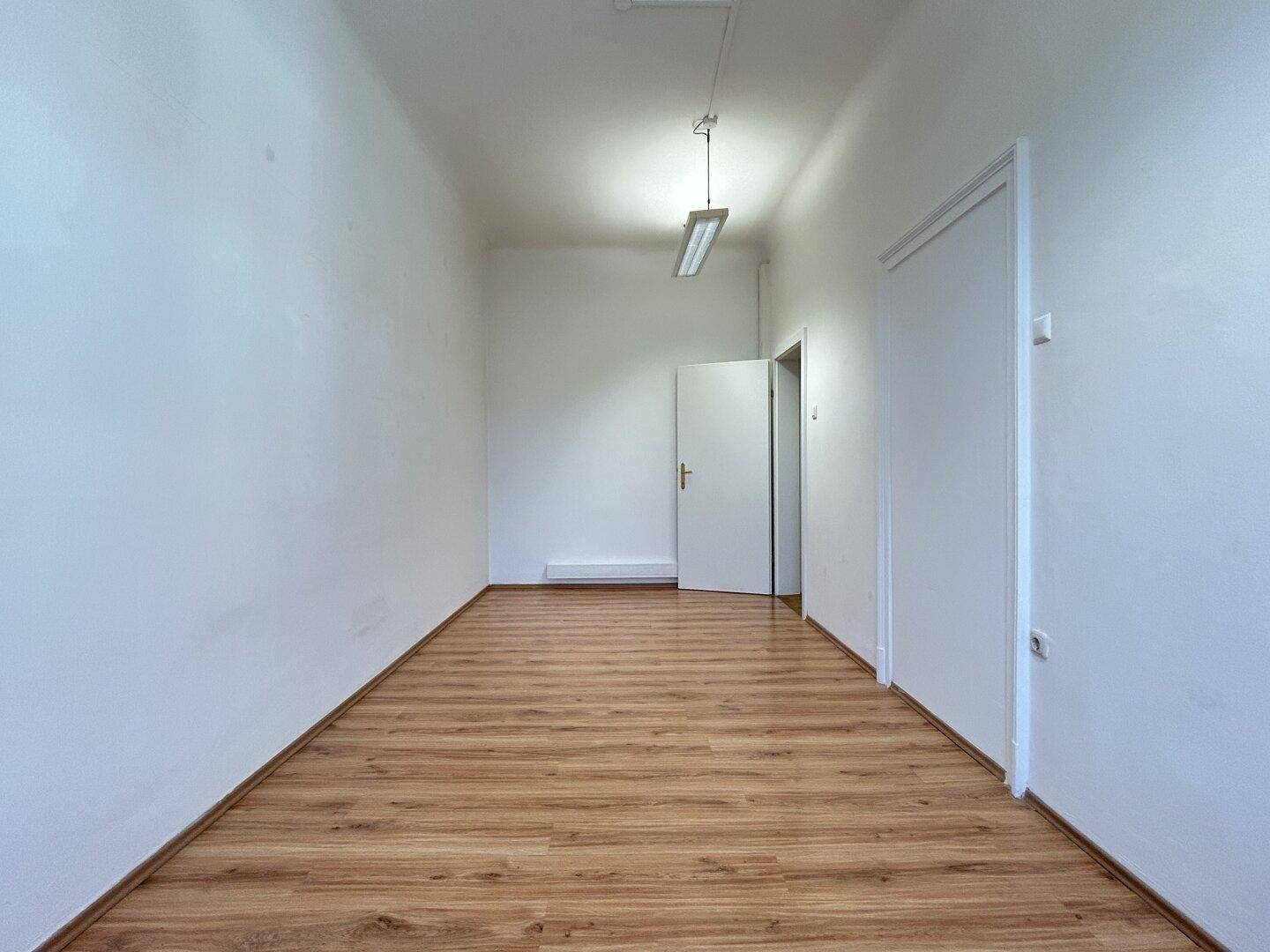 Langer, leerer Raum mit hellem Laminatboden und einer Tür am Ende.