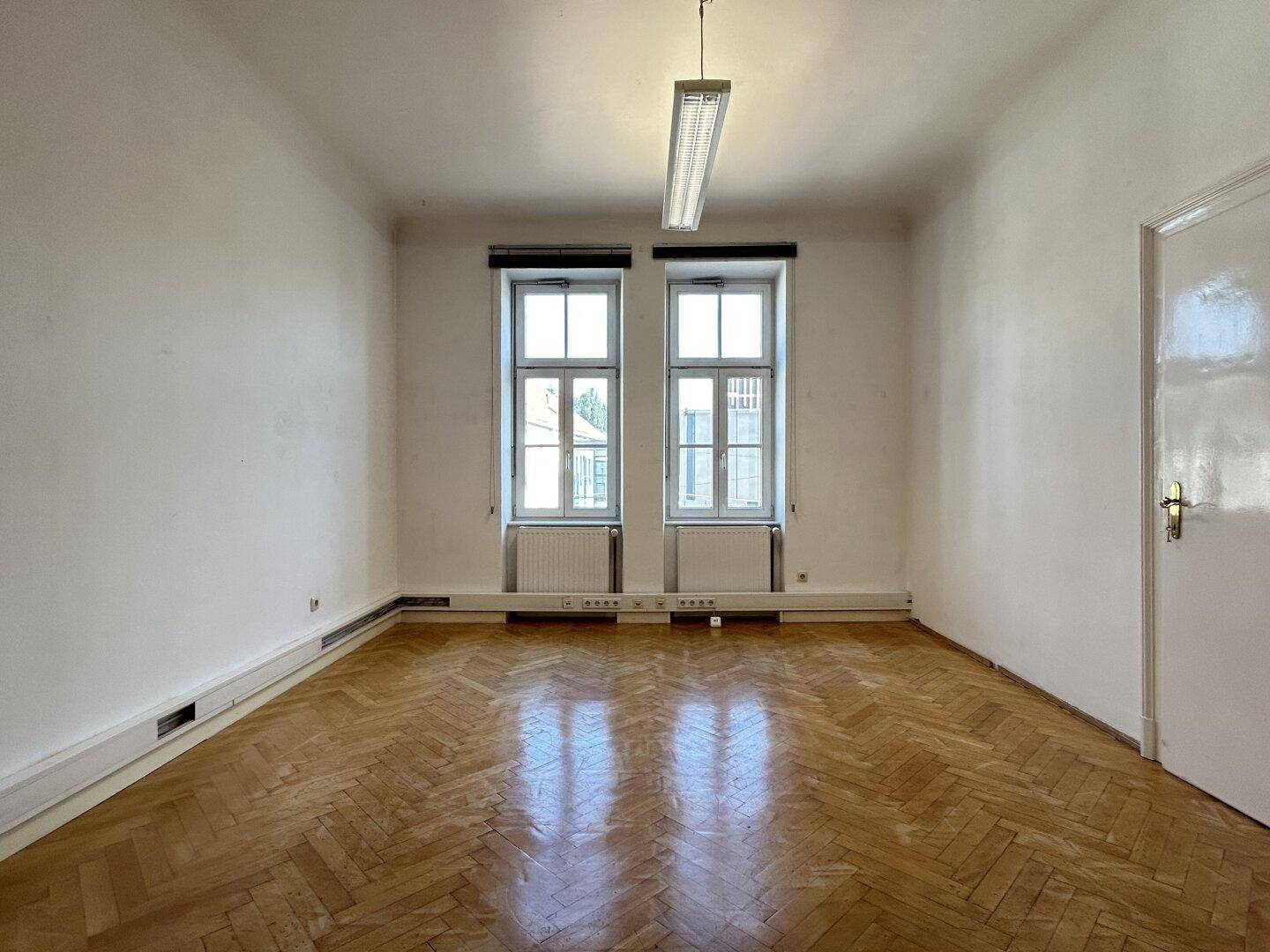 Heller Raum mit zwei Fenstern und traditionellem Fischgrätparkettboden.