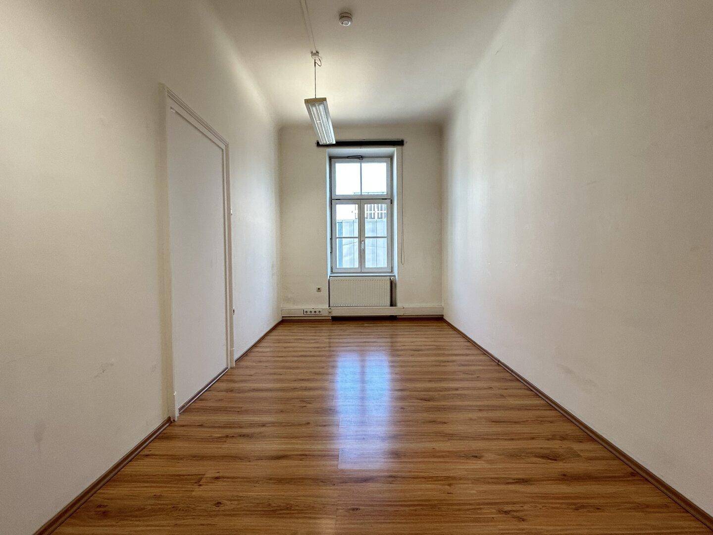 Leerer Raum mit Laminatboden und einem Fenster, das Tageslicht hereinlässt.