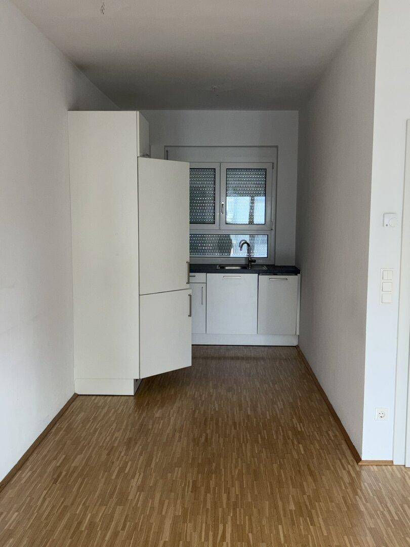 Schmale Küche mit weißer Küchenzeile, Kühlschrank und Fenster mit Rollläden.