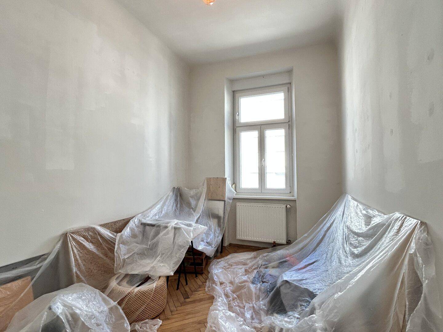 Heller Raum mit Fenster, weiß gestrichenen Wänden und abgedeckten Möbeln.
