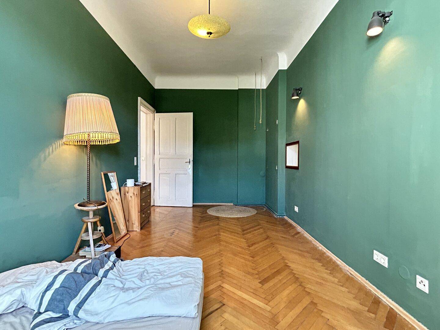 Geräumiges Schlafzimmer mit grünen Wänden, Fischgrätparkett und einer weißen Tür.