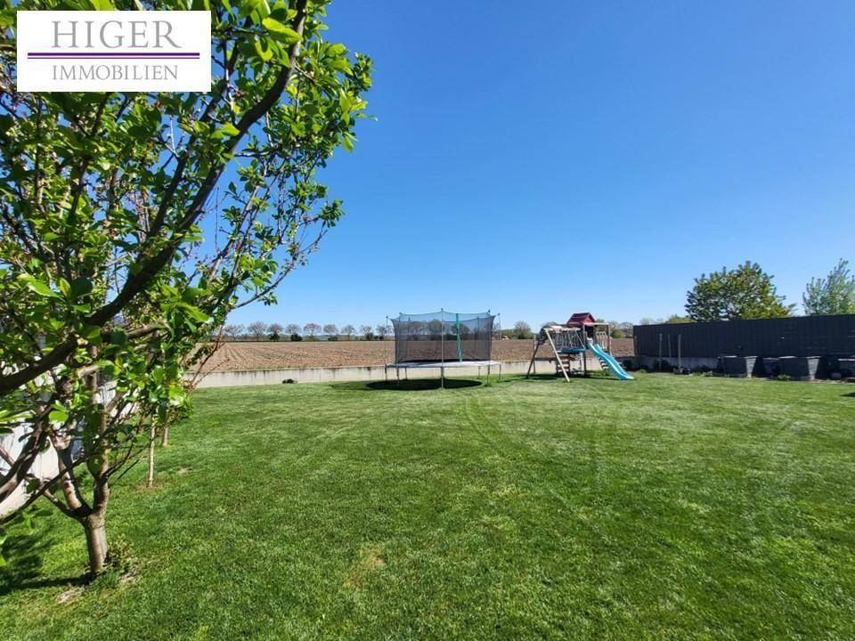 Großer Garten mit Rasenfläche, Trampolin und Spielplatz, umgeben von Feldern.